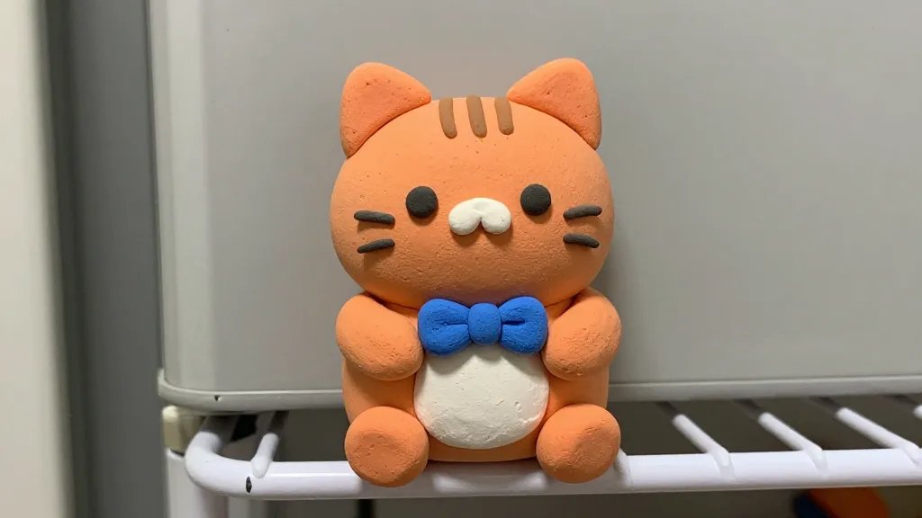 Close-up of a single clay magnet character—a chubby orange cat with tiny ears and whiskers—holding a small blue bow, perched on a metal fridge shelf with a visible magnet bond, warm lighting, detailed clay seams and tiny pores to emphasize handmade craft, no text.