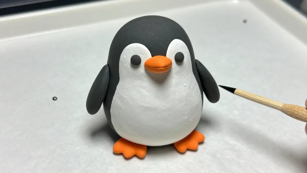 A close-up shot of a chubby penguin sculpture in progress, showing the flattened white belly and black head, with a bright orange beak and feet. Display a dotting tool and a toothpick nearby, and a tiny eye bead resting on the tray. The scene is lit to emphasize the clay textures and a subtle shine from a gloss glaze, against a clean, parchment-covered surface.