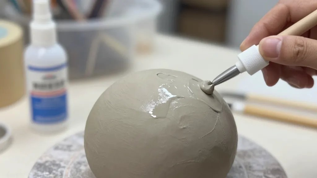 A close-up scene of a handmade air-dry clay sculpture being sealed in a well-lit studio, with a passerby-friendly gloss finish showing a subtle shine, a soft-focus background of painting supplies and a spray sealer bottle, and a clear hand applying a protective coat evenly across the clay surface.