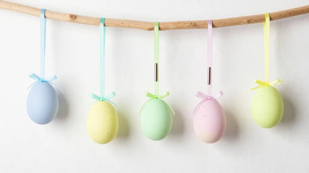 A stylish display of finished hanging Easter ornaments in a soft pastel-to-neon gradient palette, hanging from a light wooden branch against a whitewashed wall, with subtle reflections and shadows, include delicate ribbons and a few metallic pens and glitter specks in the scene, image should feel whimsical and modern, no text.
