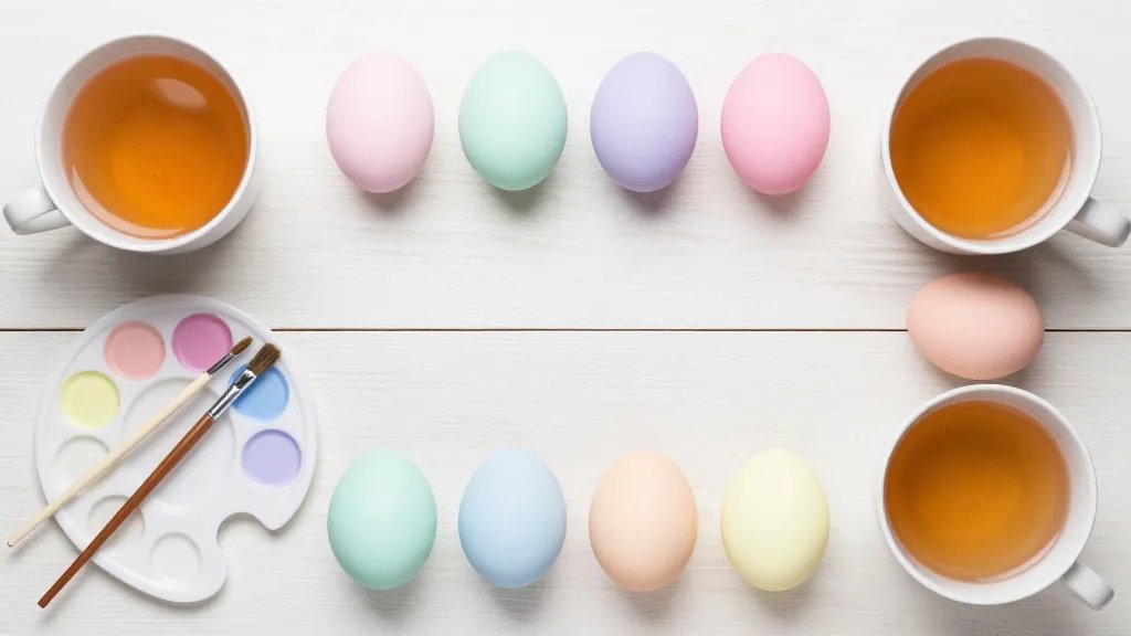 An overhead flat lay of a DIY Easter egg decorating setup: cups of tea, a small paint palette with pastel colors, brushes, and eggs in pastel hues arranged on a light wooden surface. Emphasize cohesive color harmony (pale pink, mint, lavender, baby blue, pale peach, buttercream yellow) with matte/satin finishes, gentle gradients, and a clean, modern studio aesthetic; no text.
